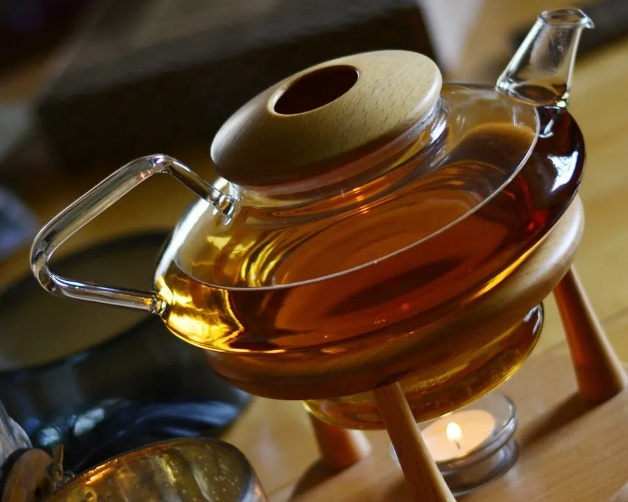 ThéiÚre en verre avec infusion chaude, posée sur un support en bois avec une bougie chauffe-plat, créant une ambiance chaleureuse et relaxante.