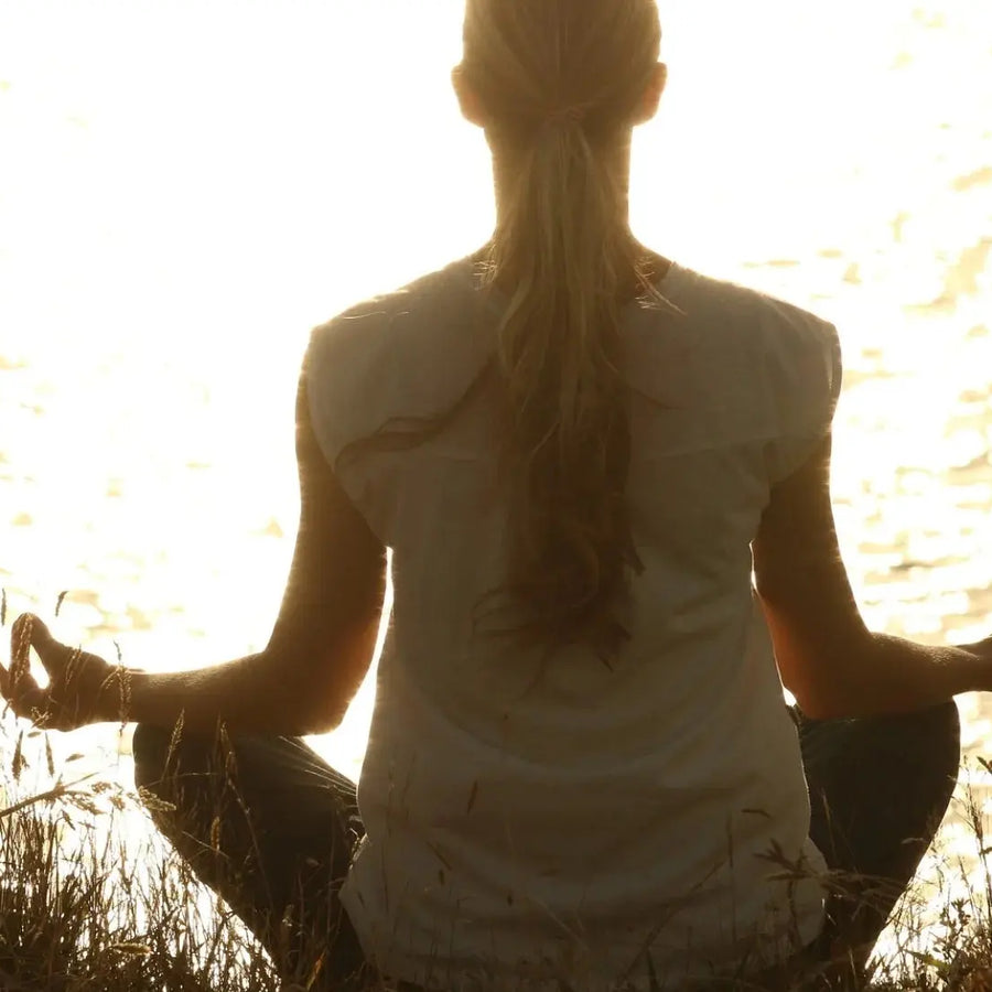 Femme méditant face au soleil couchant au bord d’un lac, en pleine nature, dans une posture de relaxation.