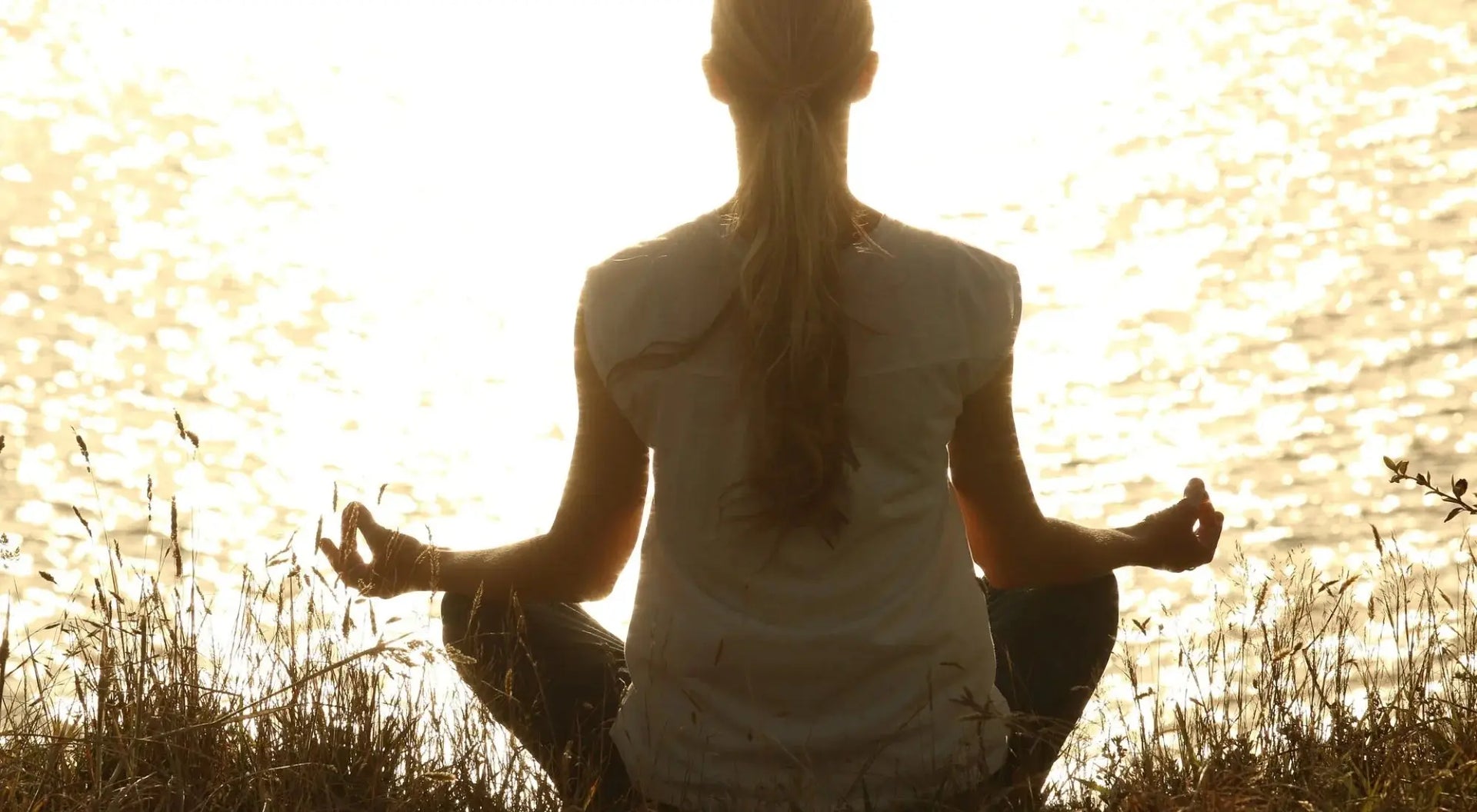 Femme méditant face au soleil couchant au bord d’un lac, en pleine nature, dans une posture de relaxation.