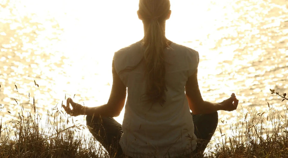 Femme méditant face au soleil couchant au bord d’un lac, en pleine nature, dans une posture de relaxation.