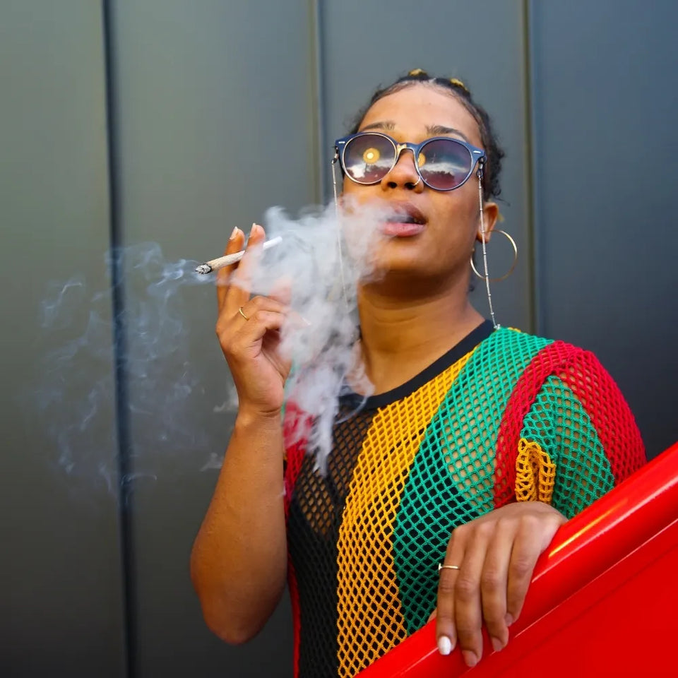 Femme stylée en tenue rasta fumant un joint de CBD devant un mur gris – format carré