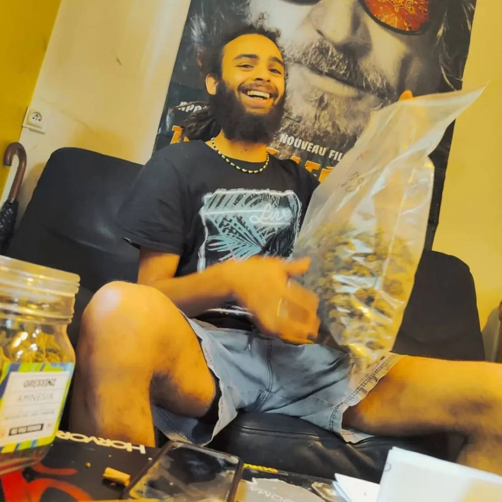 le chanteur Junior roy souriant assis sur un canapé, tenant un grand sac de fleurs de CBD, avec un bocal de Gr33nz Amnesia et divers accessoires posés devant lui. Ambiance détendue.