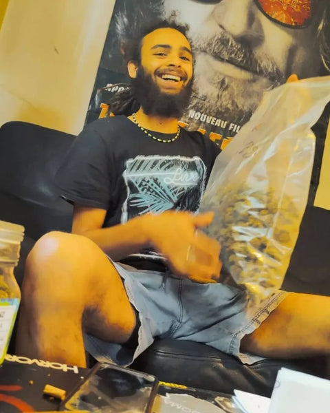 le chanteur Junior roy souriant assis sur un canapé, tenant un grand sac de fleurs de CBD, avec un bocal de Gr33nz Amnesia et divers accessoires posés devant lui. Ambiance détendue.