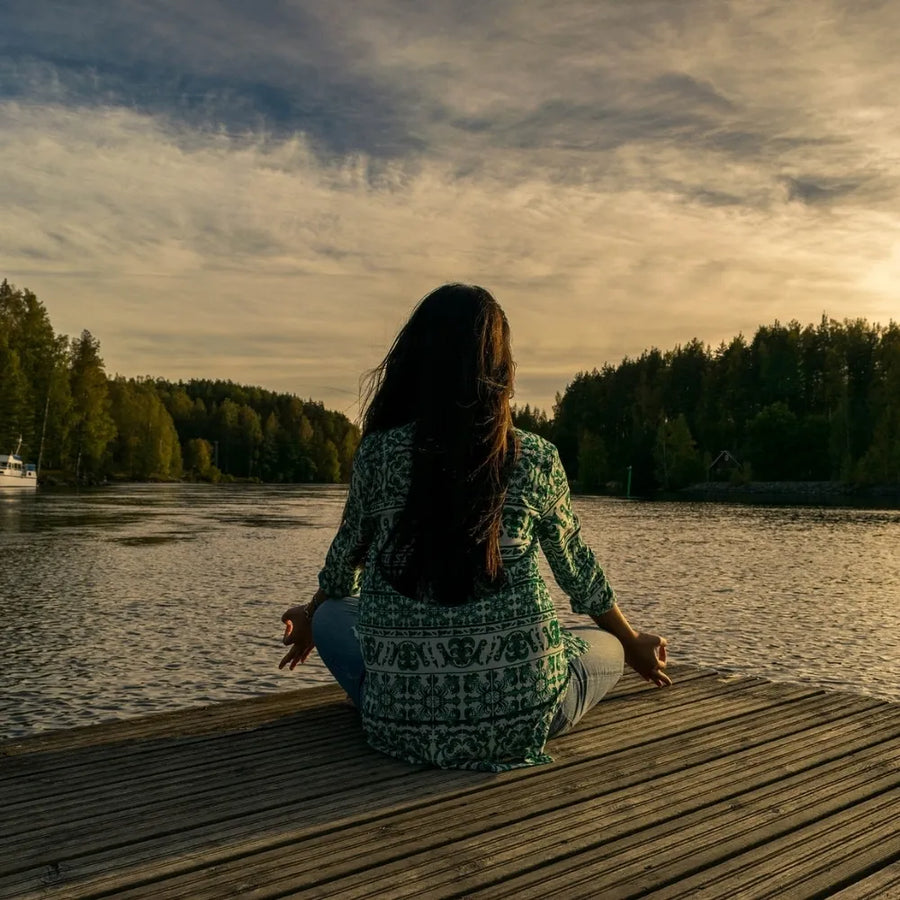 Une personne assise en méditation sur un ponton, face à un lac et entourée d'une nature paisible, au coucher du soleil.